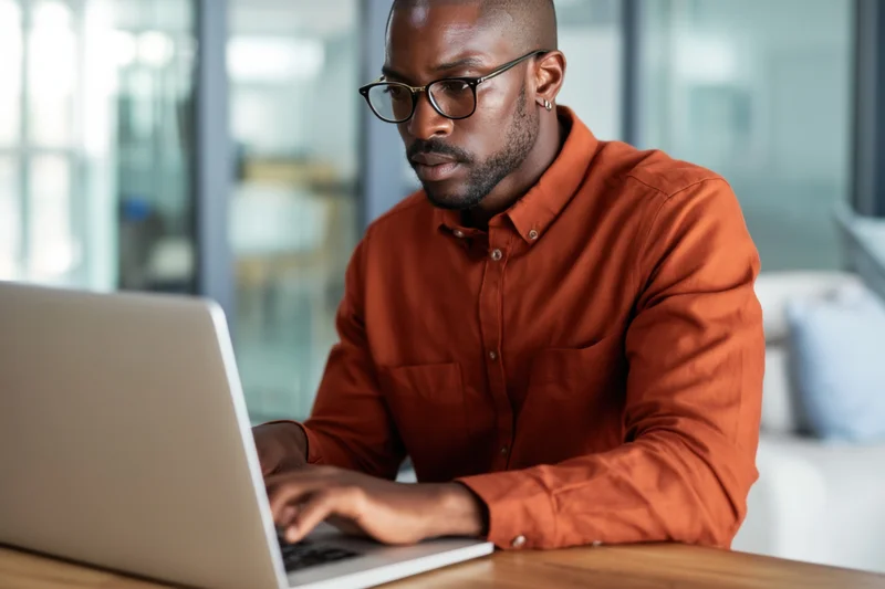 Man in rust-colored shirt typing on laptop at desk in modern office