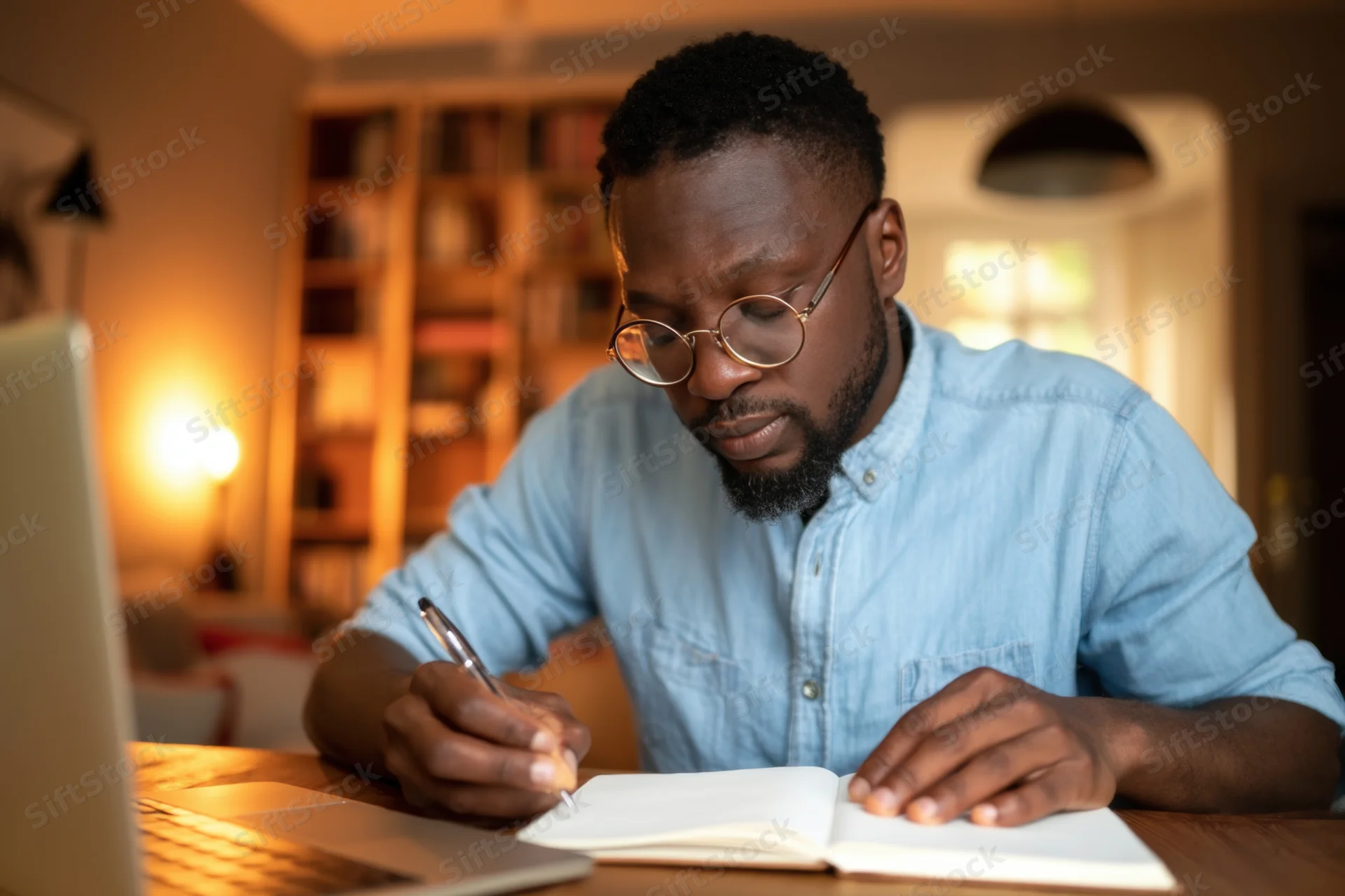 Man writing in notebook at desk beside laptop in home office setting