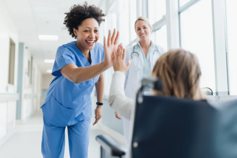 Nurse in blue scrubs high-fiving a patient in a wheelchair in a hospital corridor with a doctor in the background