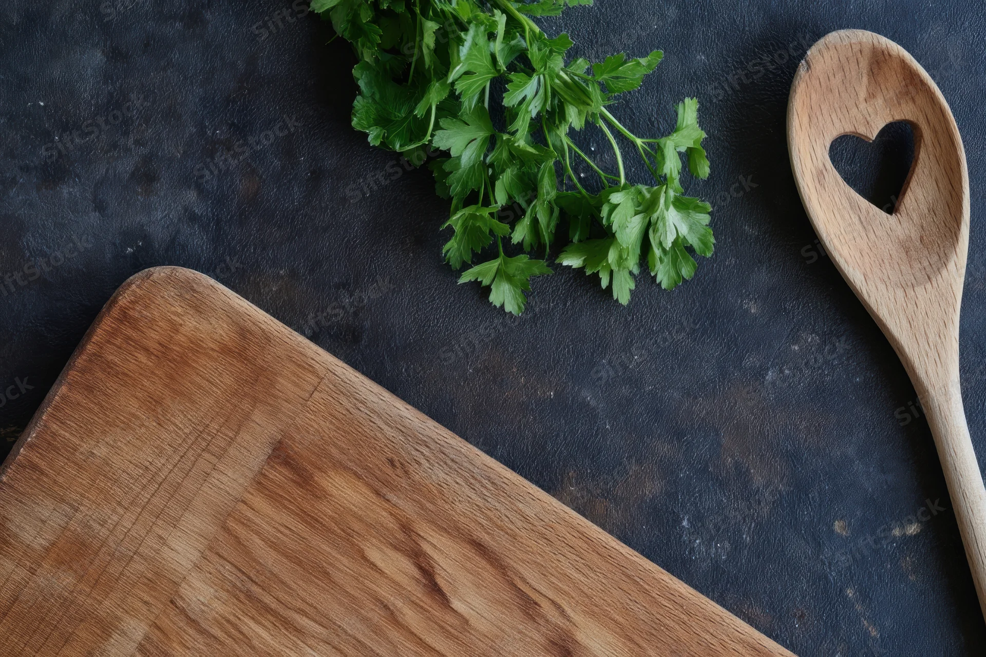 Fresh parsley and wooden spoon with heart-shaped handle on dark textured surface beside cutting board