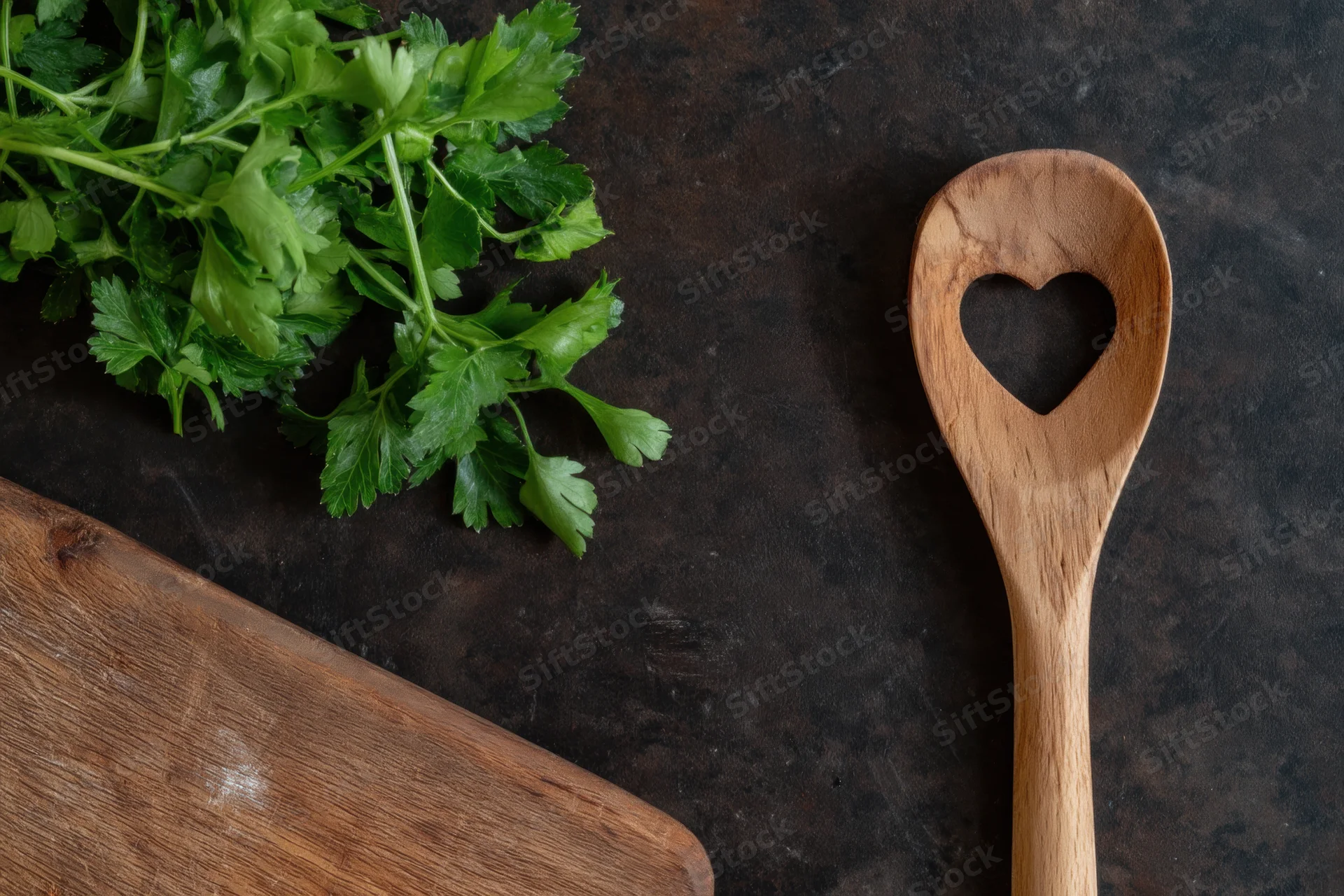 Fresh parsley and a heart-shaped wooden spoon on a dark textured surface with a cutting board, top view