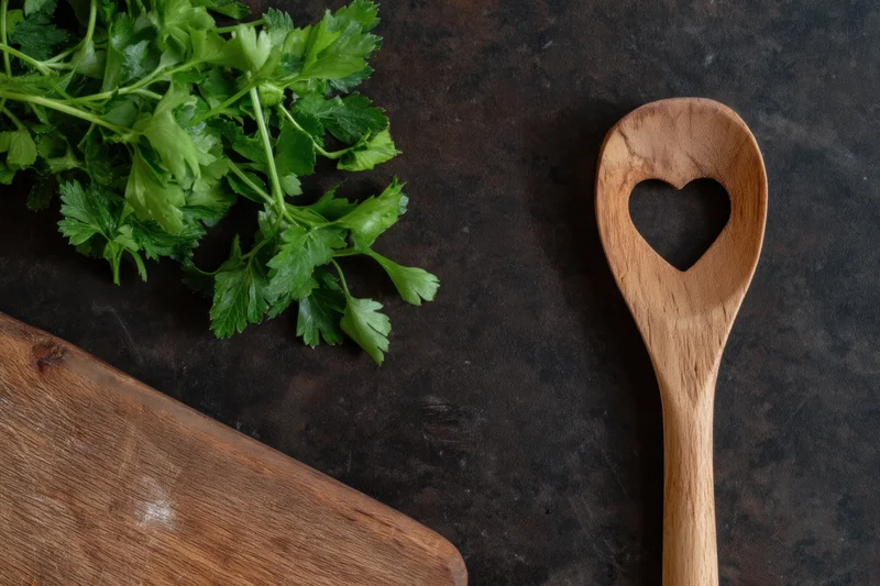 Fresh parsley and a heart-shaped wooden spoon on a dark textured surface with a cutting board, top view