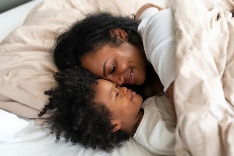 Mother cuddling with her young child on a bed, both sleeping peacefully under a blanket