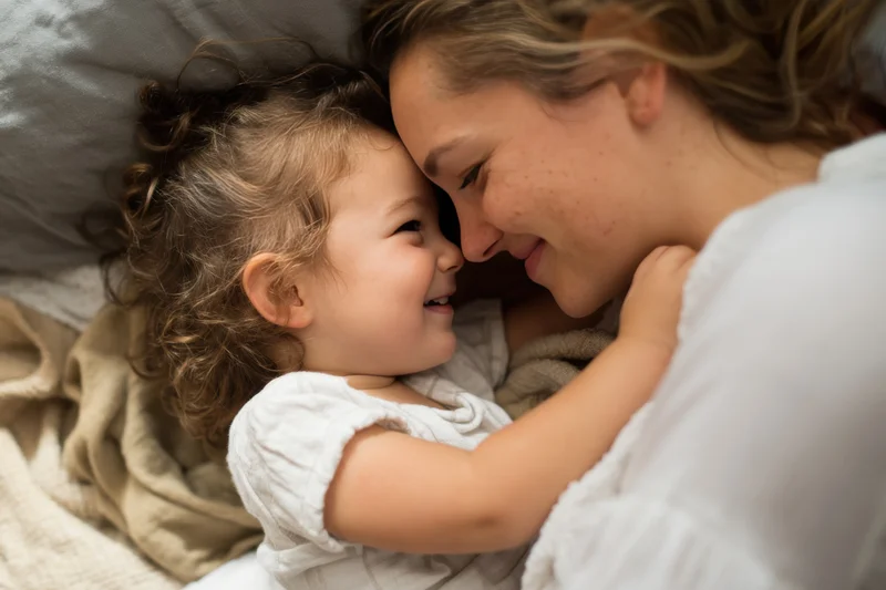 Mother and toddler cuddling closely on a bed, faces touching, smiling, wrapped in soft blankets