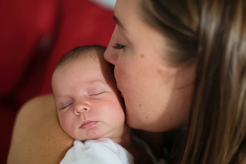 Mother kissing newborn baby held close with eyes closed