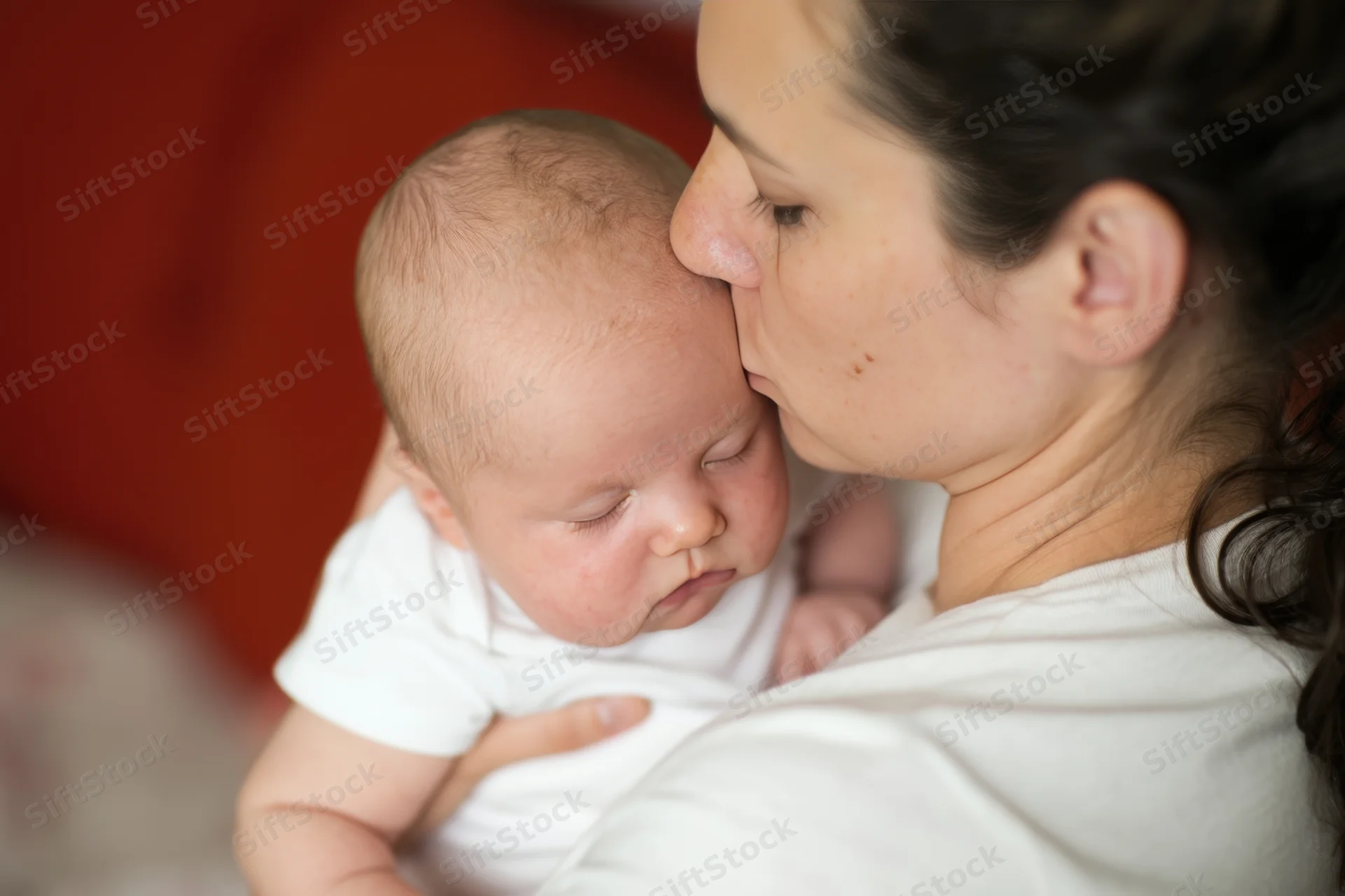 Mother kissing newborn baby held close with eyes closed indoors