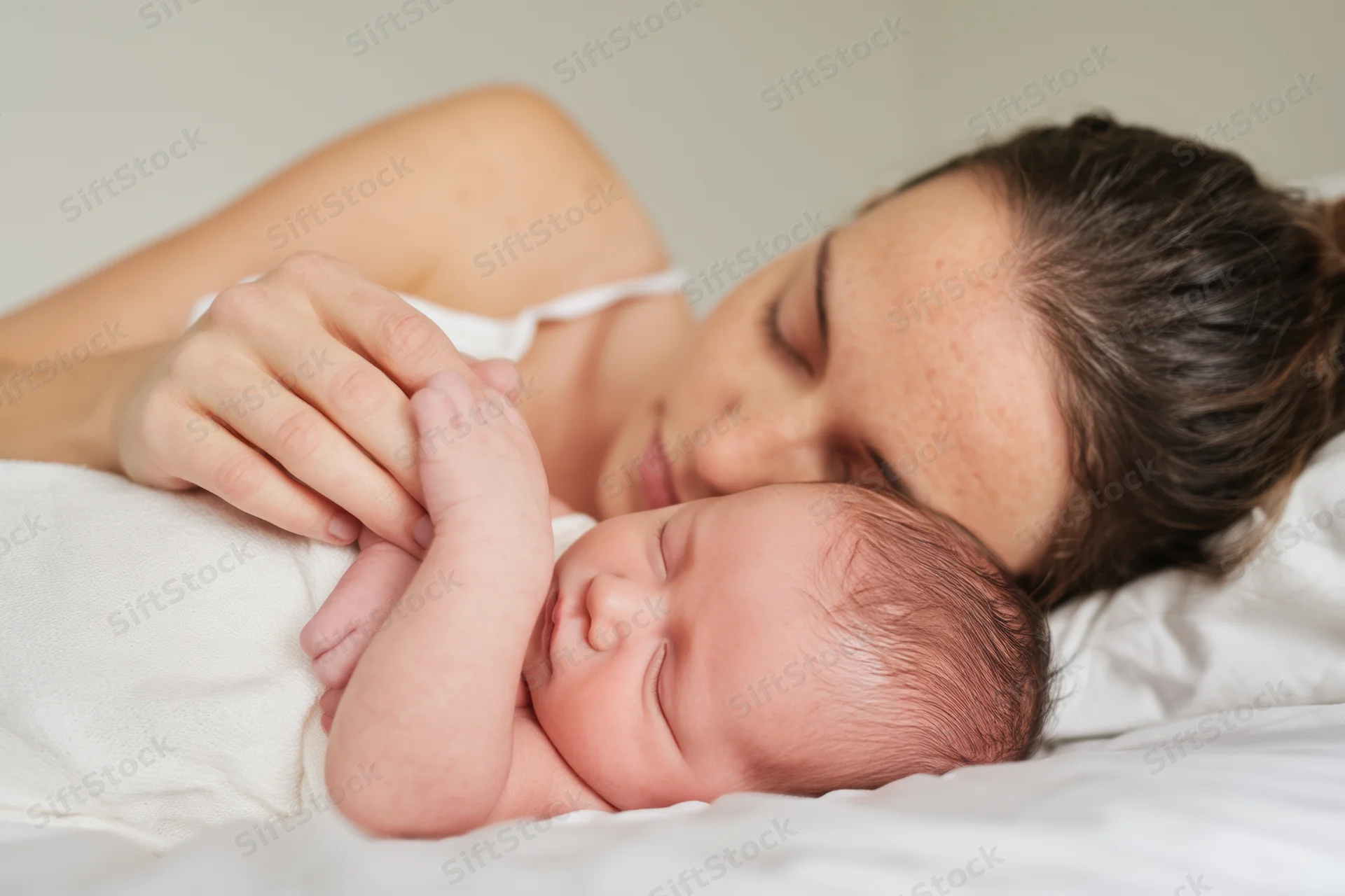 Mother lying close to sleeping newborn on white bed, gently holding baby's hand
