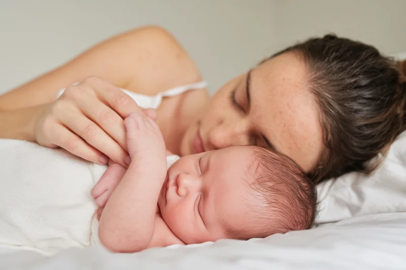 Mother lying close to sleeping newborn on white bed, gently holding baby's hand