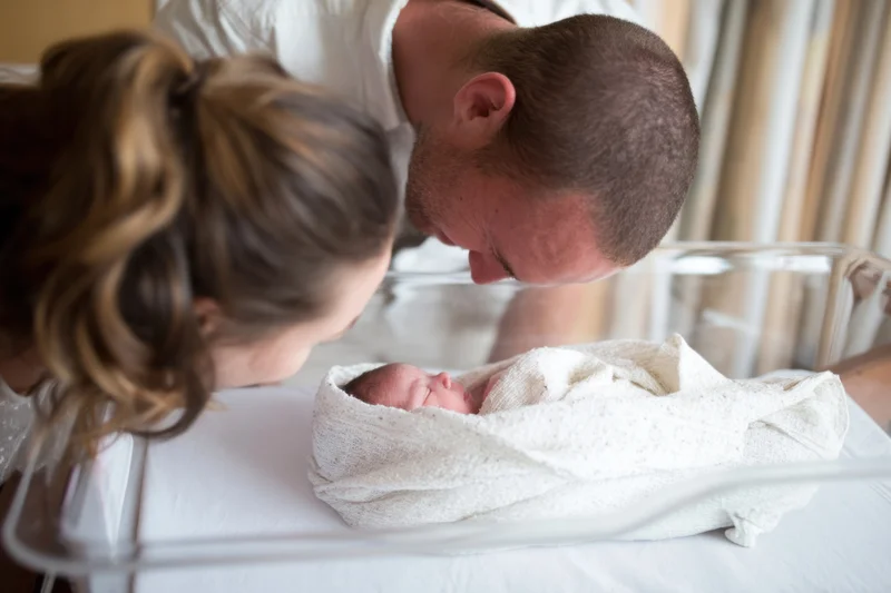 New parents leaning over their swaddled newborn in a hospital bassinet