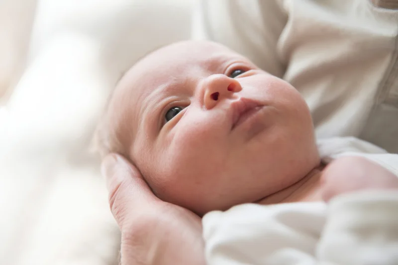 Newborn baby cradled in adult hands with eyes open indoors