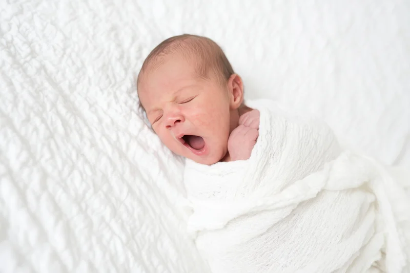 Newborn baby yawning while swaddled in a white blanket on a textured surface