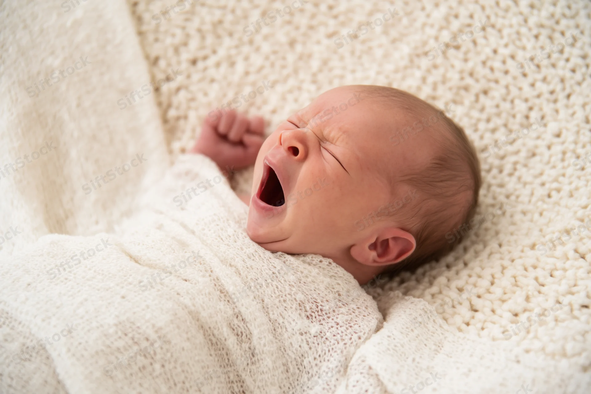 Newborn baby yawning while swaddled in a cream knitted blanket indoors