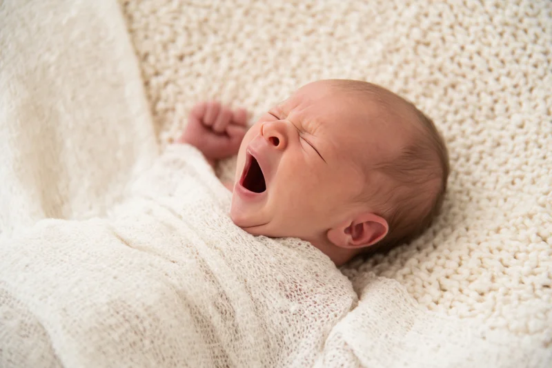 Newborn baby yawning while swaddled in a cream knitted blanket indoors