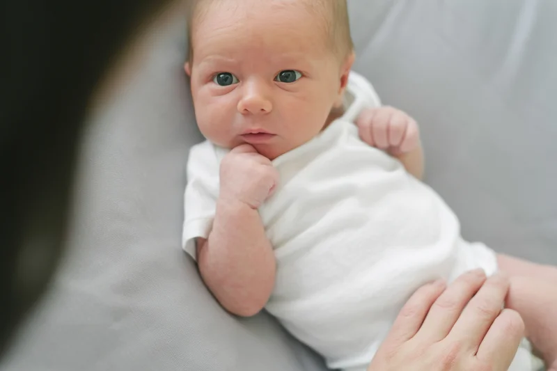 Newborn baby lying on soft gray surface in white onesie, thumb in mouth, hand resting nearby