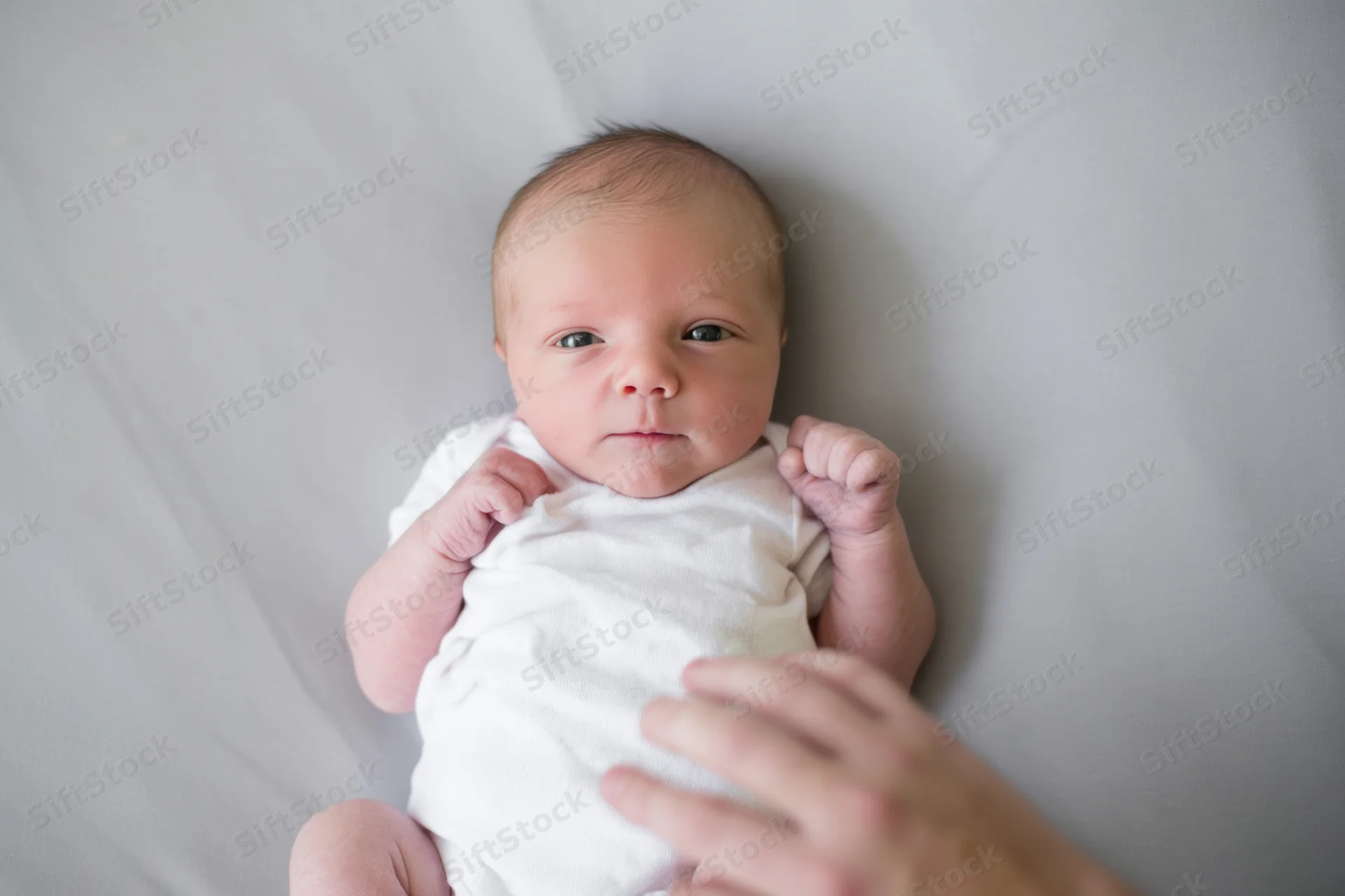 Newborn baby lying on gray surface in white onesie, overhead view with hand reaching toward it