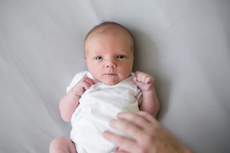 Newborn baby lying on gray surface in white onesie, overhead view with hand reaching toward it