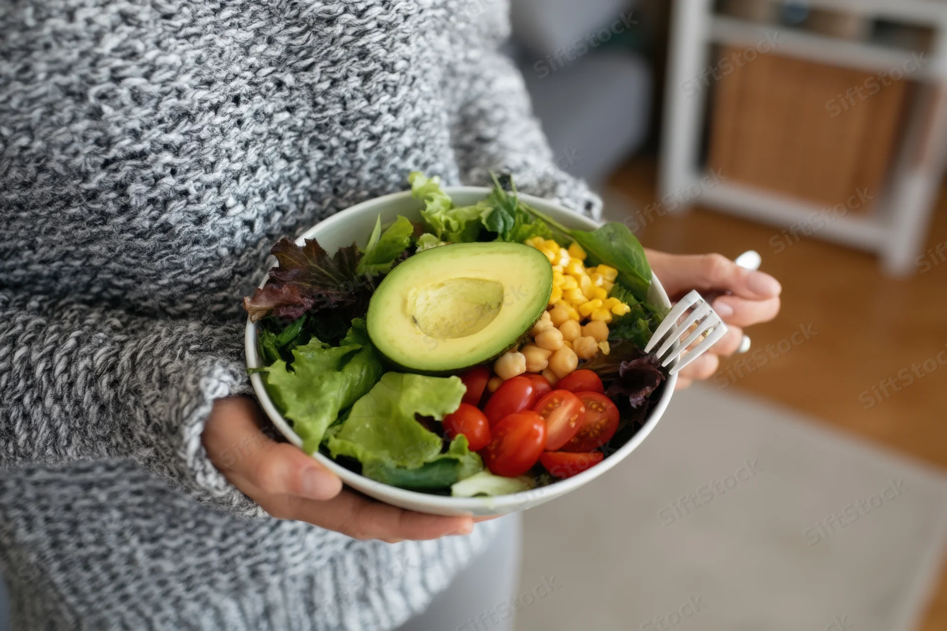 Woman holding a bowl of salad with avocado, tomatoes, corn and chickpeas in a home kitchen setting