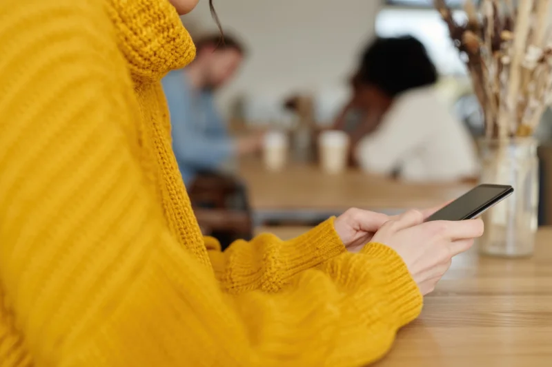 Woman in yellow knit sweater using smartphone at wooden cafe table
