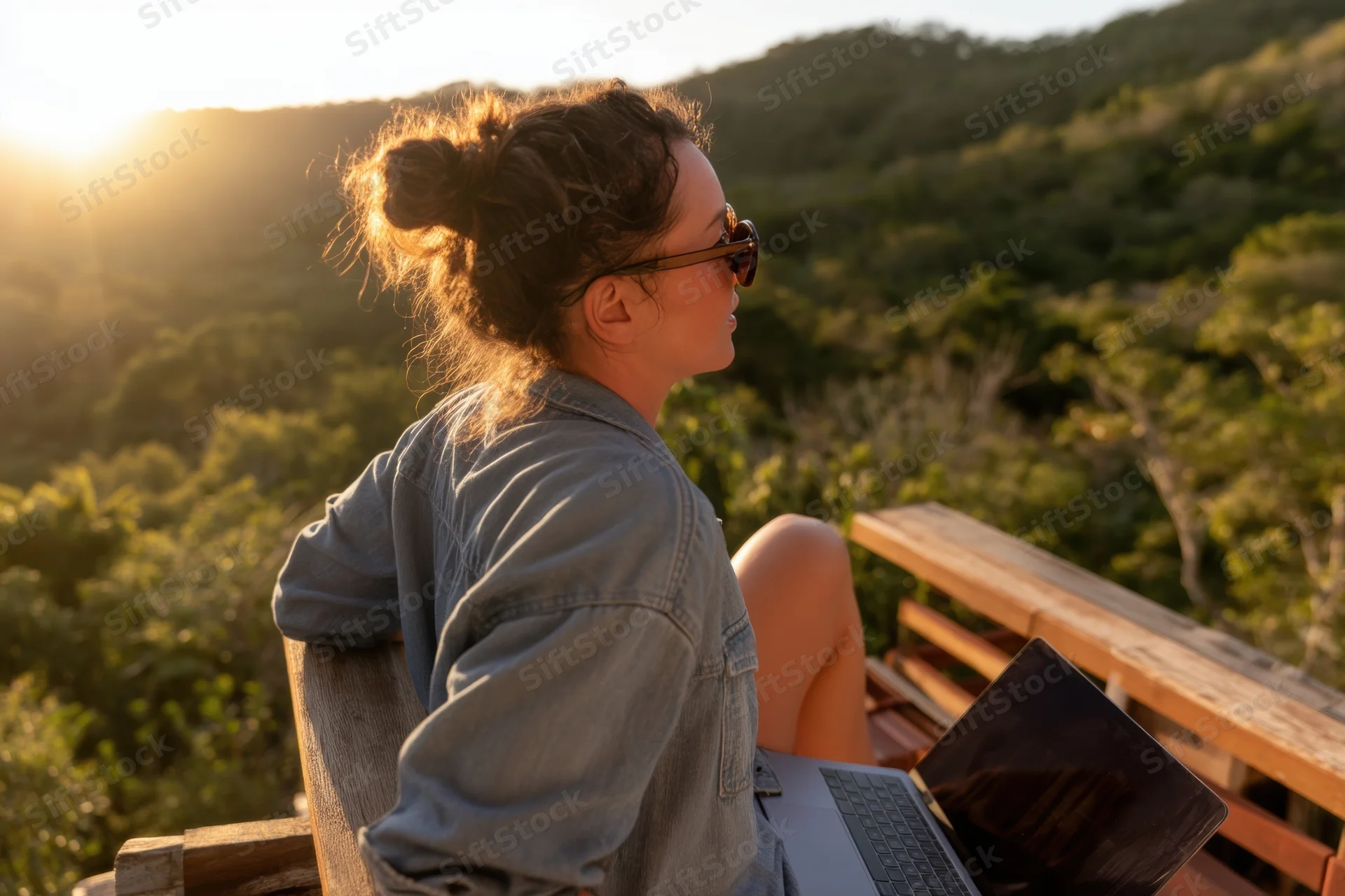 Woman sitting on wooden balcony with laptop, overlooking forested hills at sunset