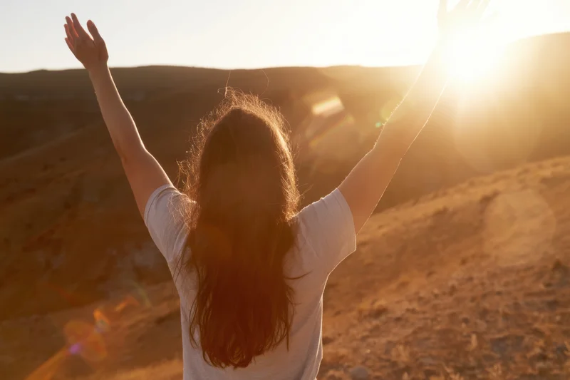 Woman with arms raised on sunlit hillside at sunset