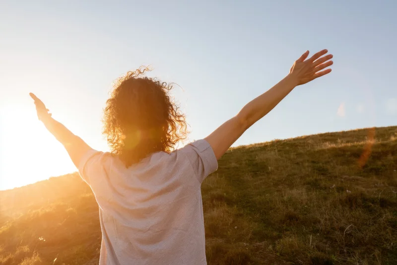 Woman with arms raised facing sunset on a grassy hillside