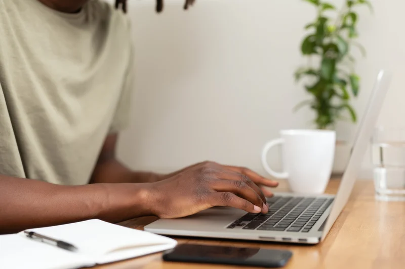 Person typing on laptop at wooden desk with notebook and smartphone nearby