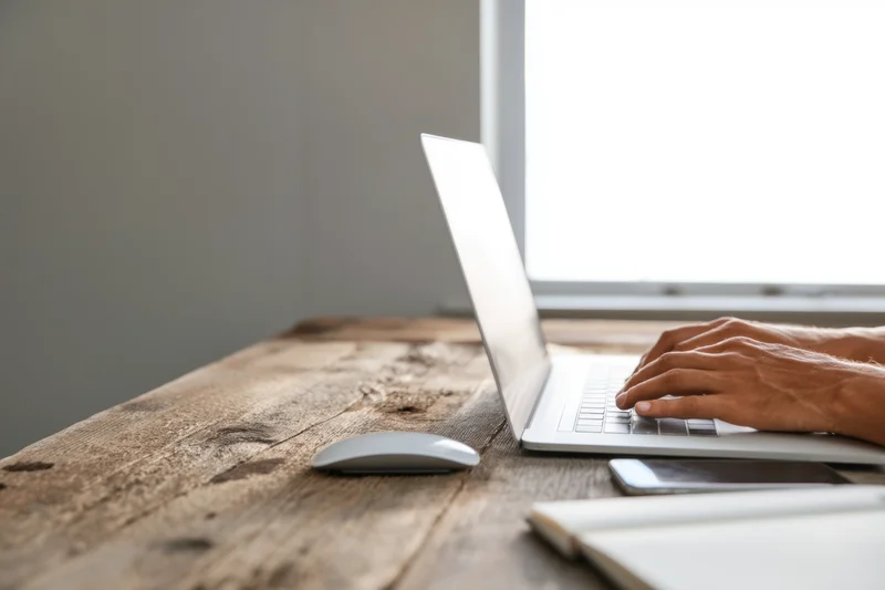 Hands typing on a silver laptop at a rustic wooden desk by a window