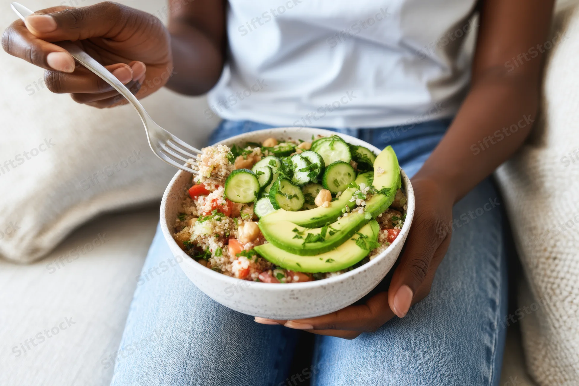 Woman eating a quinoa salad with avocado and cucumber from a ceramic bowl while sitting on a couch