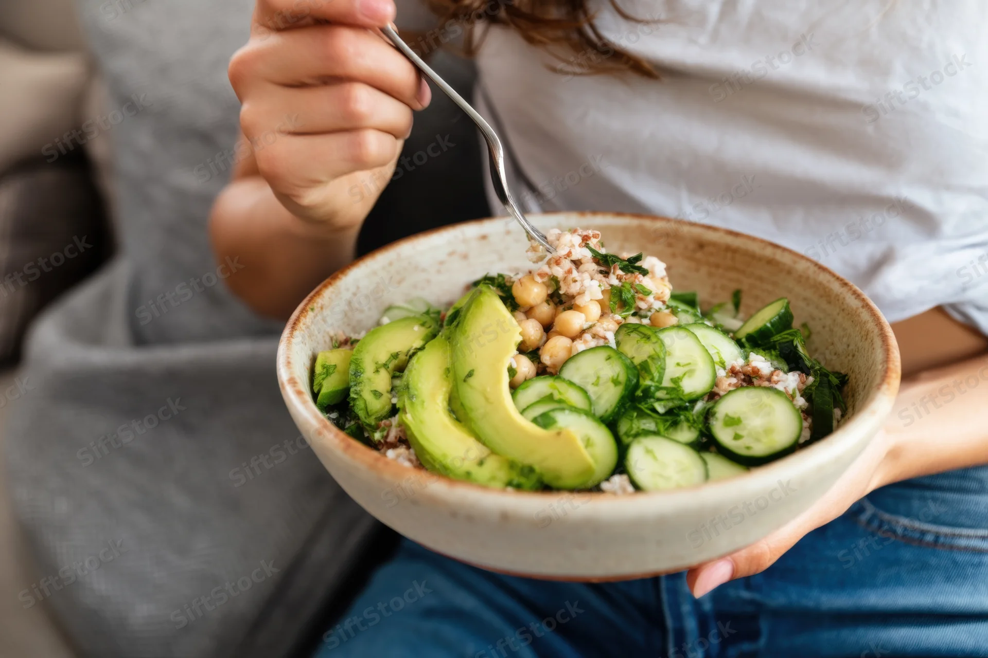 Woman eating a salad with avocado and cucumber from a ceramic bowl while sitting on a couch