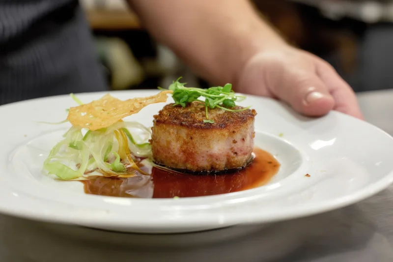 Seared meat medallion with sauce and garnish on a white plate in a kitchen setting
