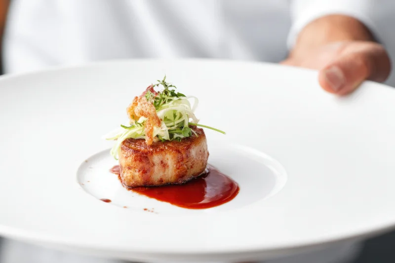 Chef presenting a seared meat medallion with red sauce and herb garnish on a white plate