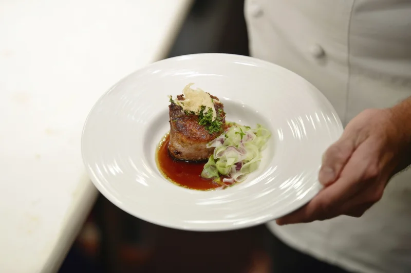 Chef holding a plated seared meat medallion with sauce and garnish in a restaurant kitchen