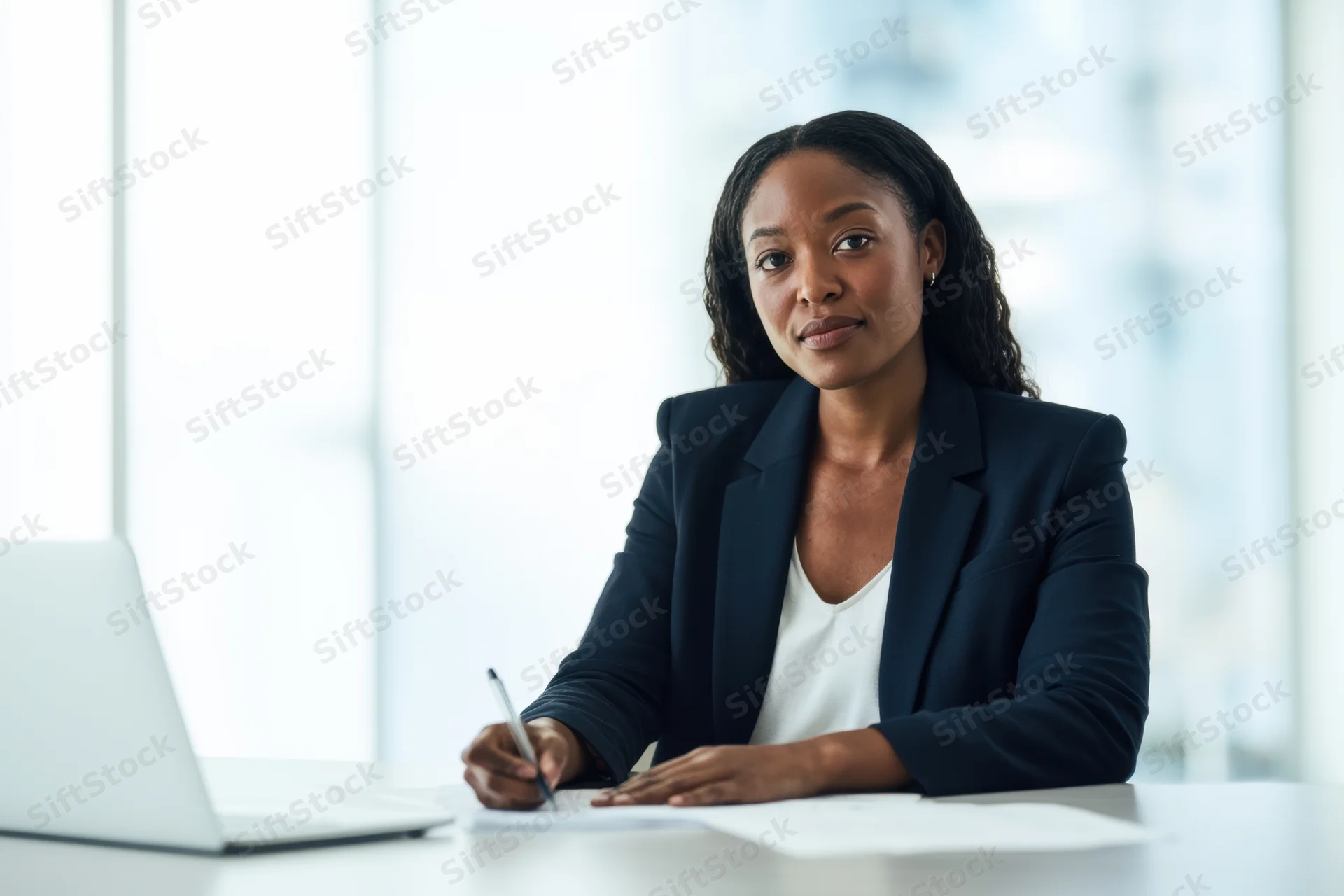 Woman in dark blazer writing at desk with laptop in office
