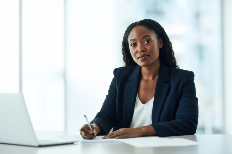 Woman in dark blazer writing at desk with laptop in office