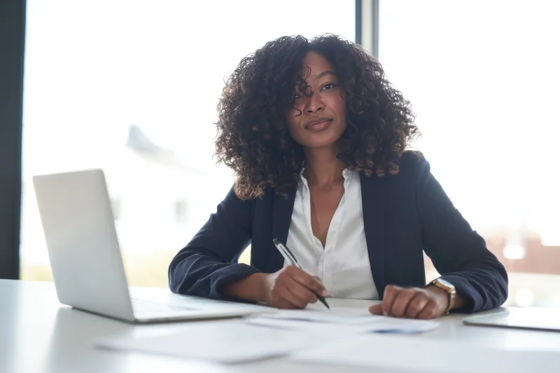 Woman in dark blazer writing with pen at desk in office near window