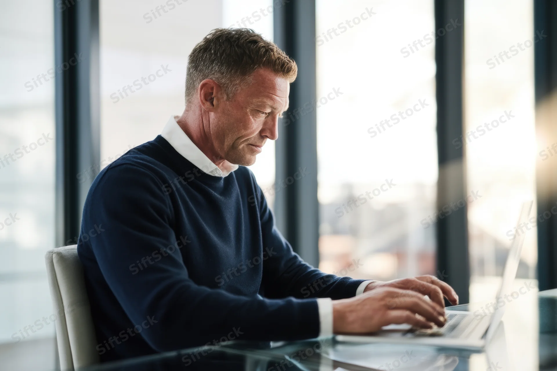 Man typing on laptop at glass desk in modern office near window