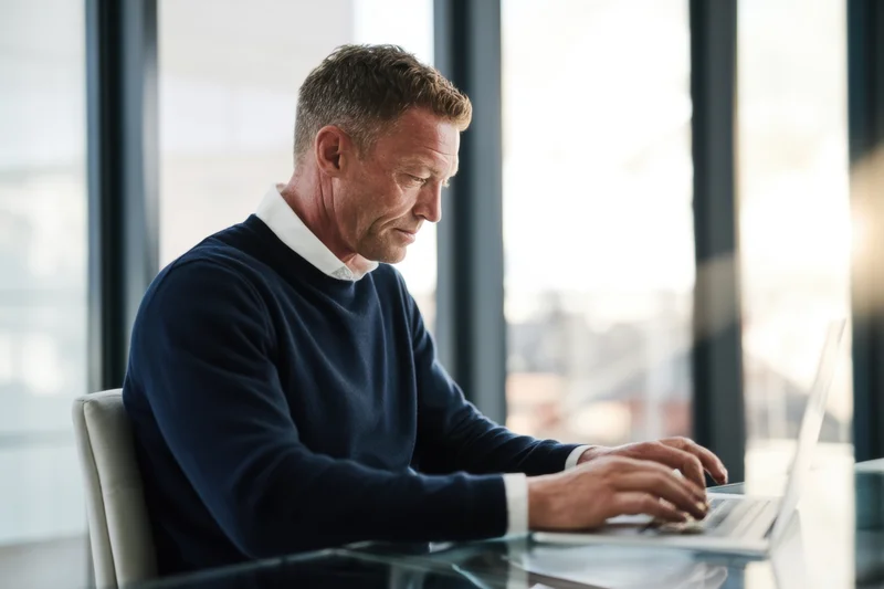 Man typing on laptop at glass desk in modern office near window