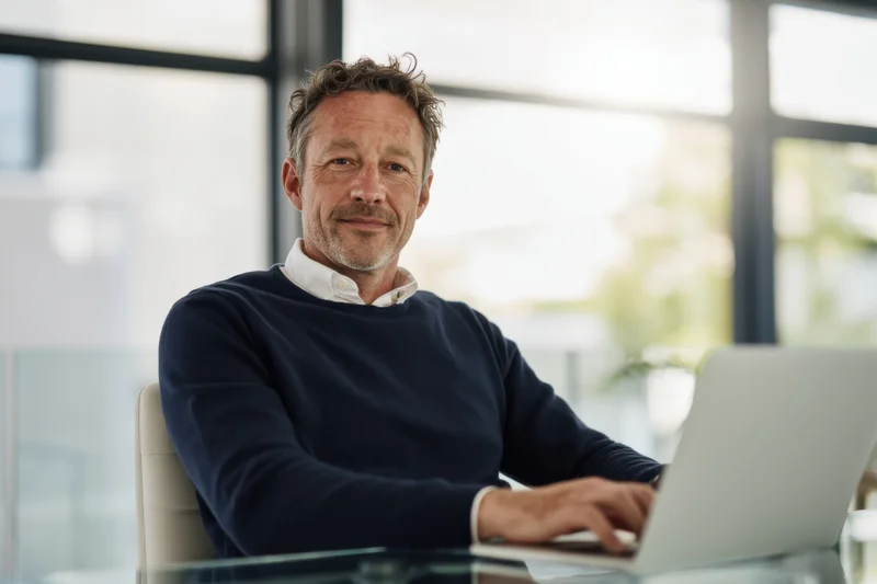 Man smiling while typing on laptop at glass desk in modern office