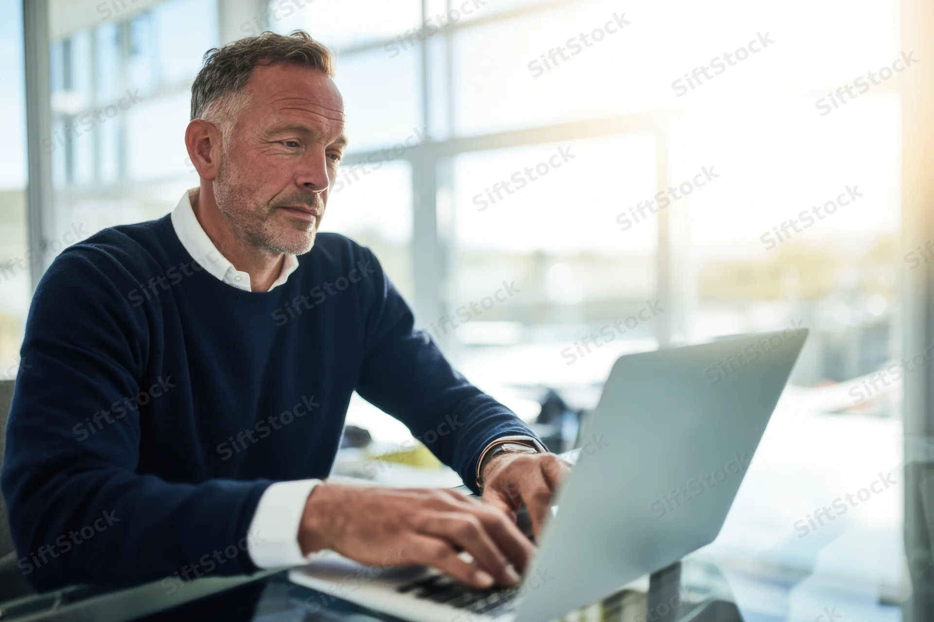 Mature man typing on laptop at office desk by window