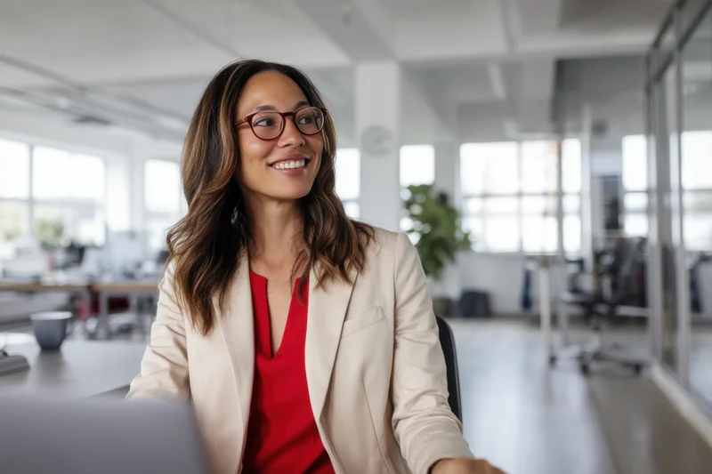 Woman in beige jacket smiling at desk in modern open-plan office