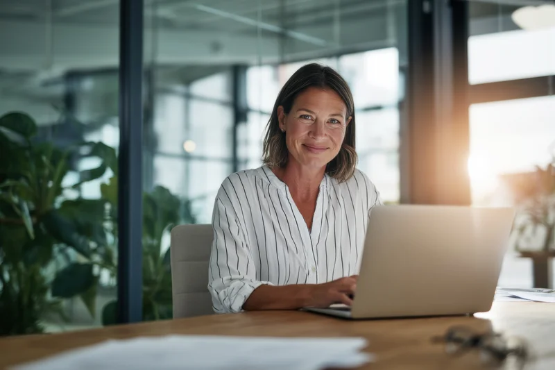 Woman smiling at camera while working on laptop at office desk