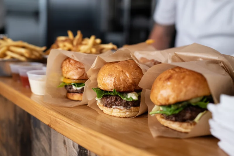 Row of burgers wrapped in parchment on wooden counter with fries and condiments in background