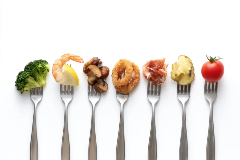 Seven forks each holding a different food item arranged in a row on white background