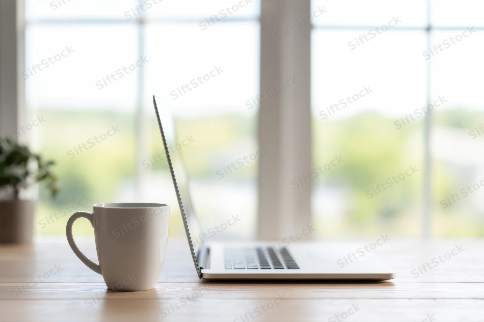 Laptop and white mug on wooden desk by window in home office workspace