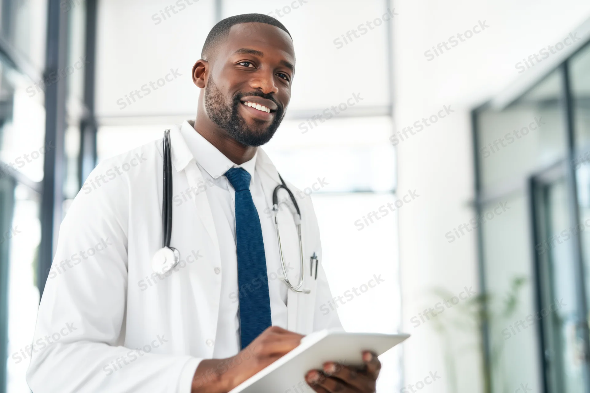 Smiling black male doctor in white lab coat holding tablet in modern clinic interior