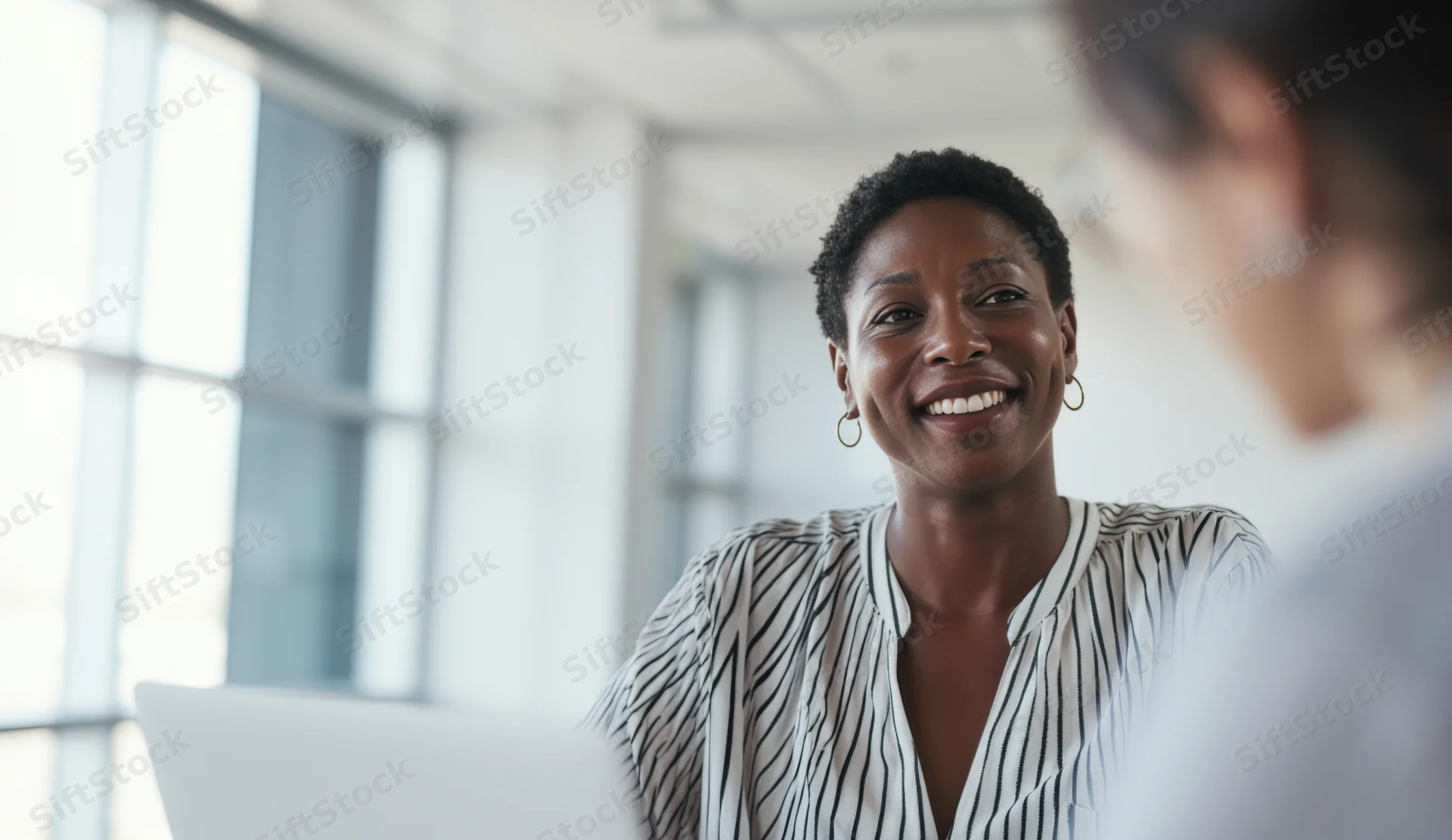 Smiling woman in striped blouse talking to colleague at office desk