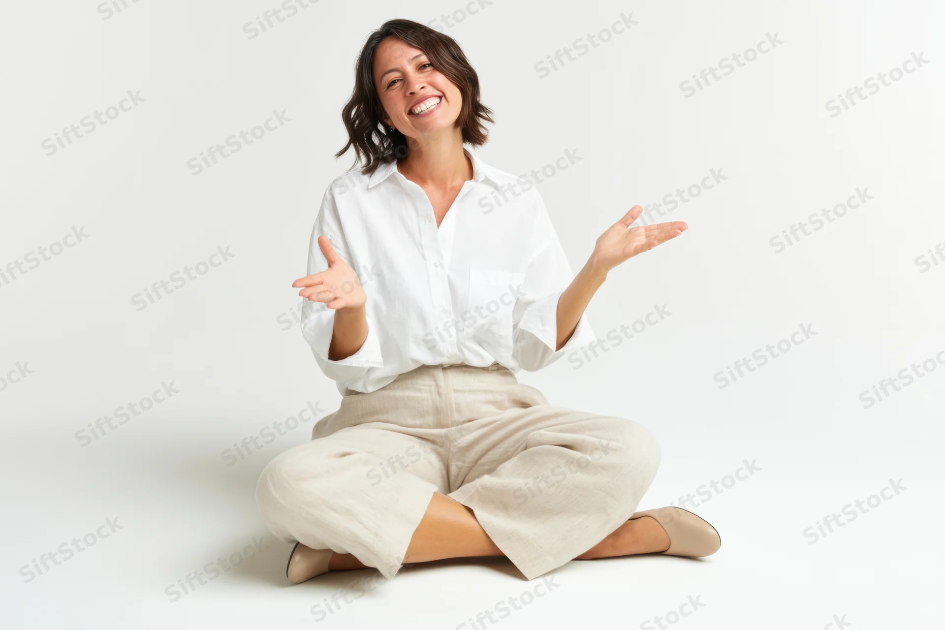 Smiling woman seated crosslegged on white studio backdrop with open hands gesturing welcomingly