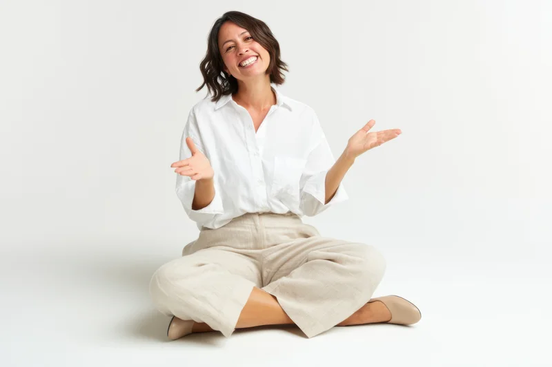 Smiling woman seated crosslegged on white studio backdrop with open hands gesturing welcomingly