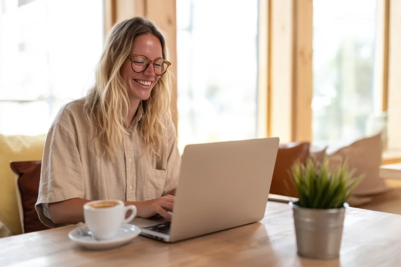 Smiling blonde woman with glasses typing on laptop at wooden table in bright cafe