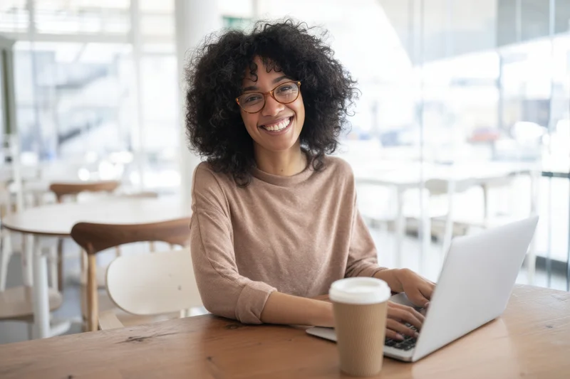 Smiling woman with curly hair and glasses typing on laptop at wooden table in bright cafe